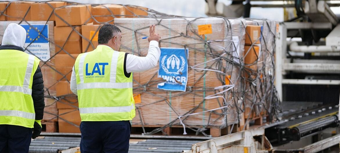 Deux travailleurs de l'aéroport vêtus de gilets de sécurité jaunes se tiennent près de palettes de fournitures de secours qui sont chargées sur un avion. La cargaison est marquée par les logos du HCR et de l'Union européenne, représentant l'aide humanitaire aux familles déplacées au Liban.