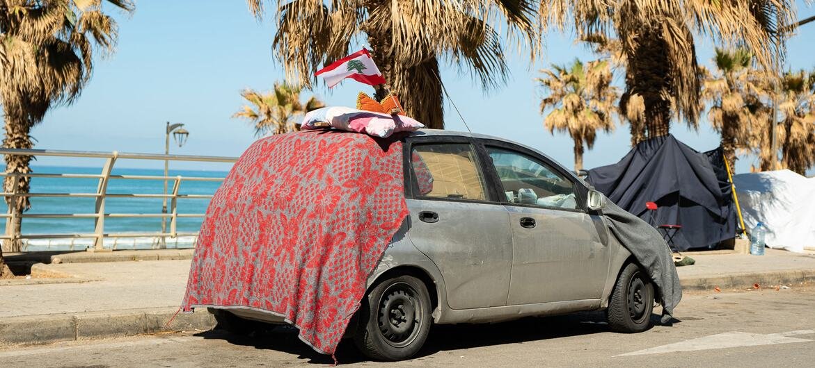 Guerre au Moyen-Orient : cibler les infrastructures civiles viole le droit international, rappelle Guterres 1 Un refuge provisoire d'une famille déplacée à Beyrouth, au Liban, construit à partir d'une voiture et de couvertures avec un drapeau libanais sur le dessus, près de tentes sur le front de mer.