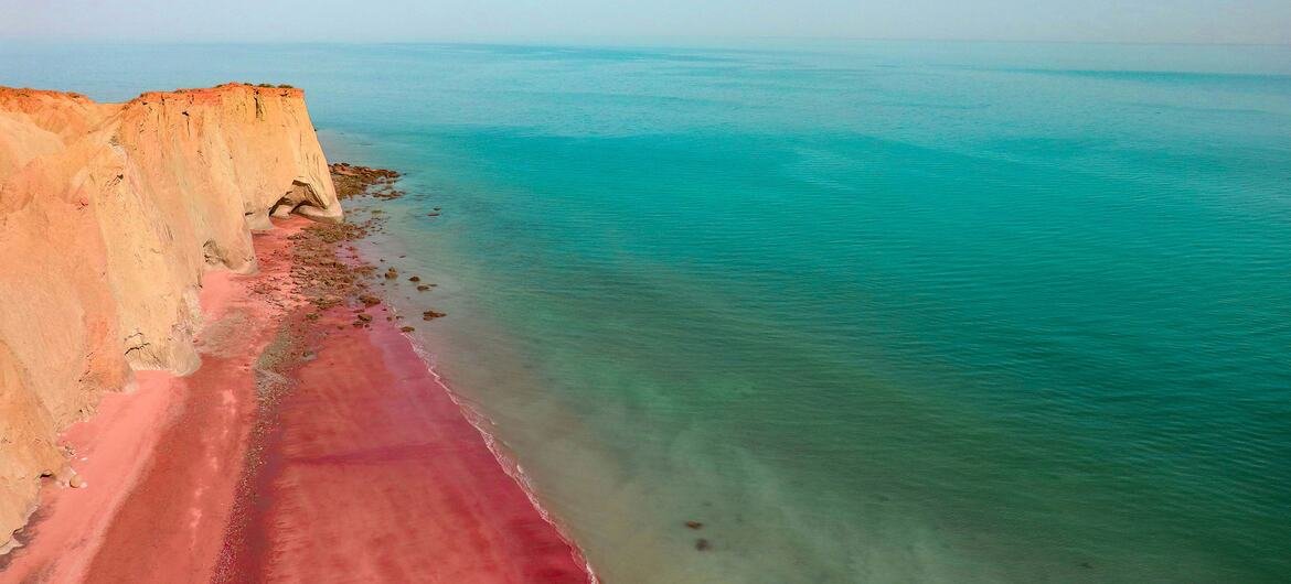 Une vue de la plage rouge sur l'île d'Ormuz en Iran, avec le littoral de sable rose distinctif et des falaises rouges rencontrant l'eau turquoise sous un ciel bleu.