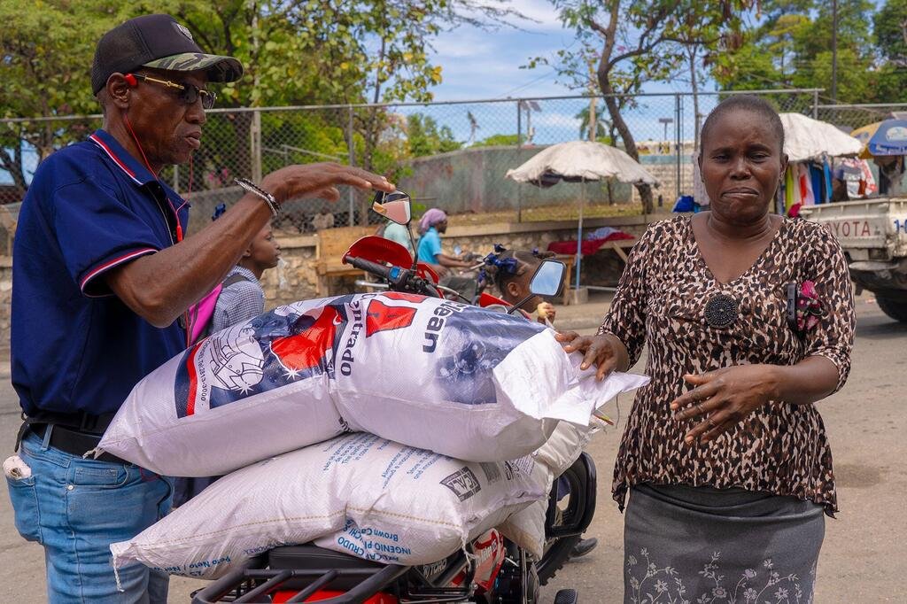 Un homme charge de grands sacs de riz sur un motocyclette pendant qu'une femme, identifiée comme Dina, observe. Cette image illustre le Programme alimentaire mondial (PAM) fournissant une aide alimentaire à Canapé Vert, en Haïti.