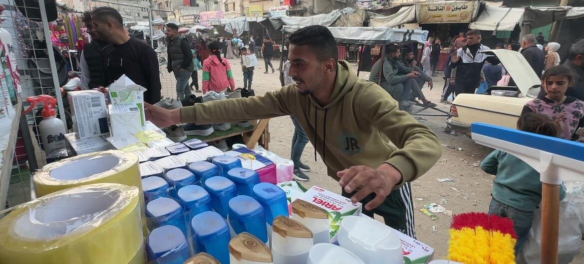Un homme palestinien vend des articles ménagers, tels que des produits d'entretien et du ruban adhésif, dans un marché de rue improvisé à Khan Younis, dans la bande de Gaza. À l'arrière-plan, des bâtiments endommagés sont visibles, illustrant l'« économie de survie » où des jeunes diplômés occupent des emplois modestes en raison du conflit en cours.