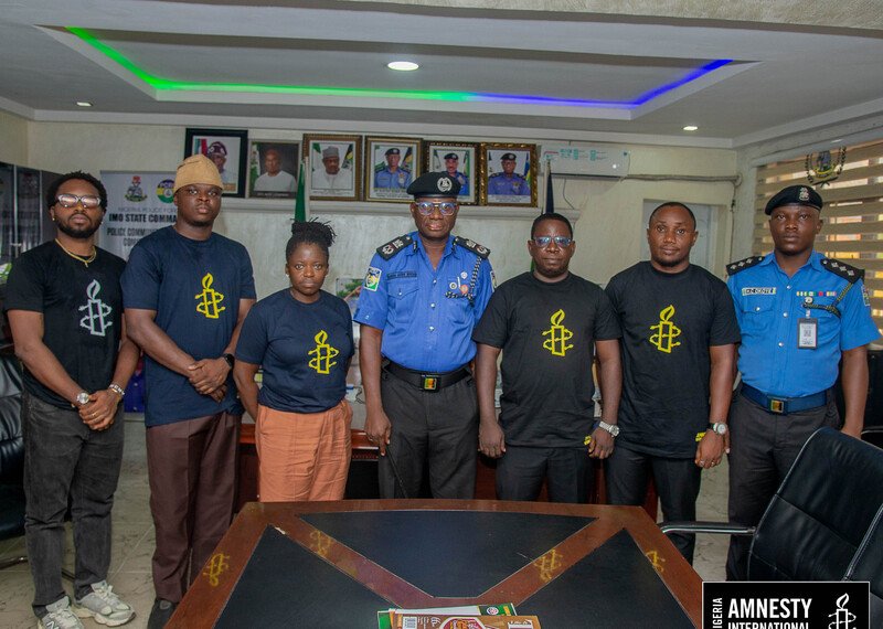 A group of people stand seriously in a dark office - some are wearing black t-shirts, while the other two wear a police uniform.