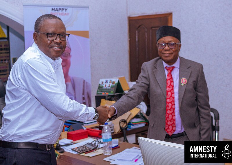 Two men shake hands over a desk, piled high with papers.