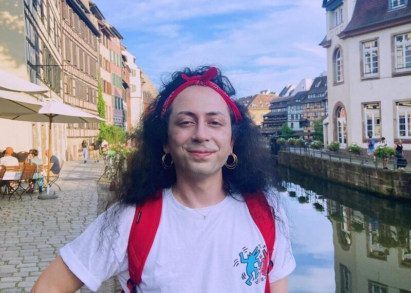 A person, wearing a red backpack and red headband, smiles happily to camera. They are stood by a beautiful canal, where people sit out on either side, drinking coffee.
