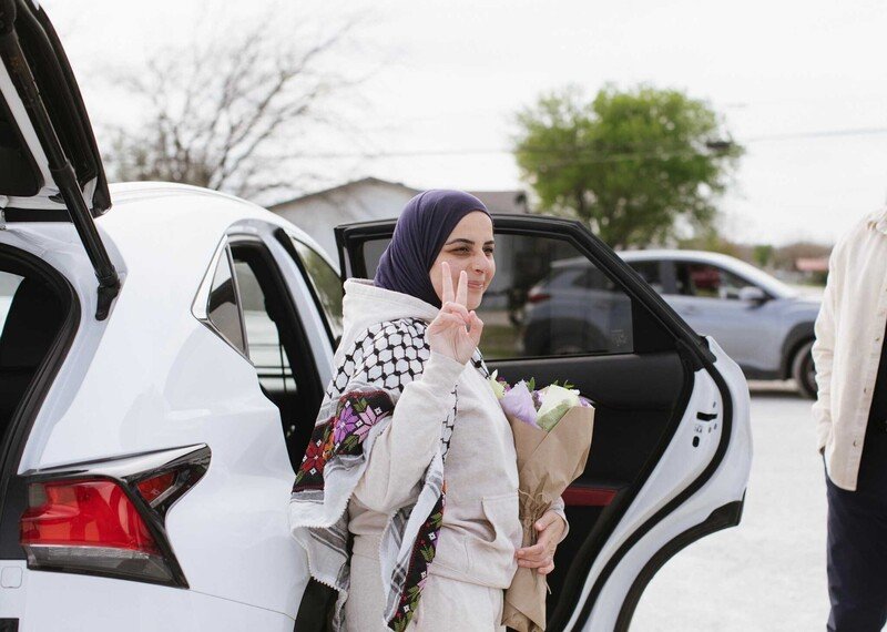 A woman in a white tracksuit and black headscarf gives the peace sign as she steps out of white car.