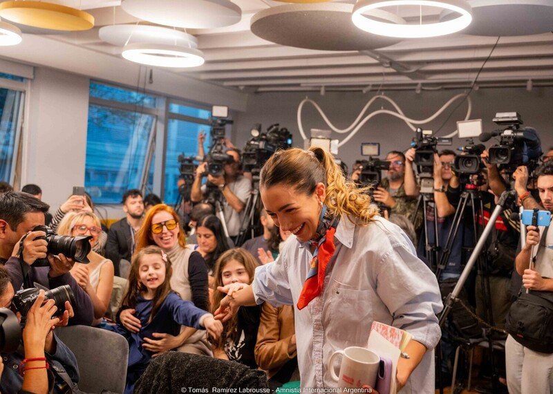 A woman is wearing a blue shirt and red neckerchief. She smiles joyfully, as she walks through a packed room of cameras and smiling people.